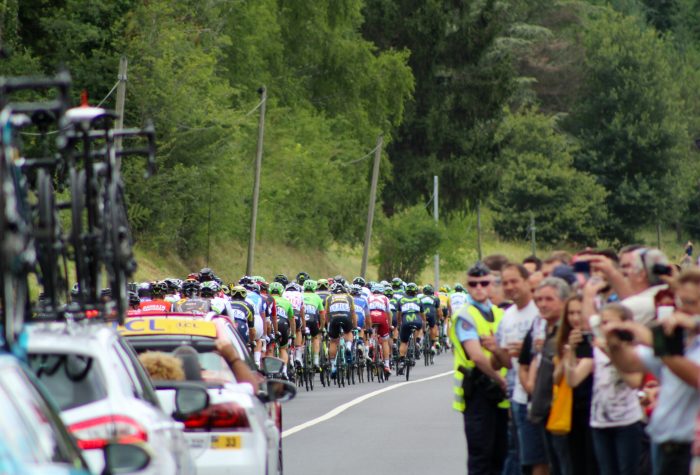A crowd take pictures of a large group of racing cyclists being escorted through a street.